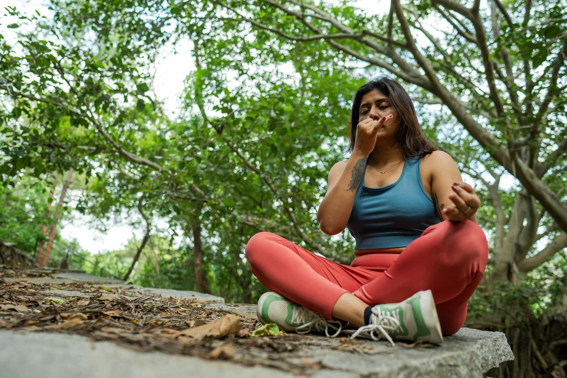 A woman sitting cross-legged outdoors under trees, practicing a breathing exercise with one hand near her nose