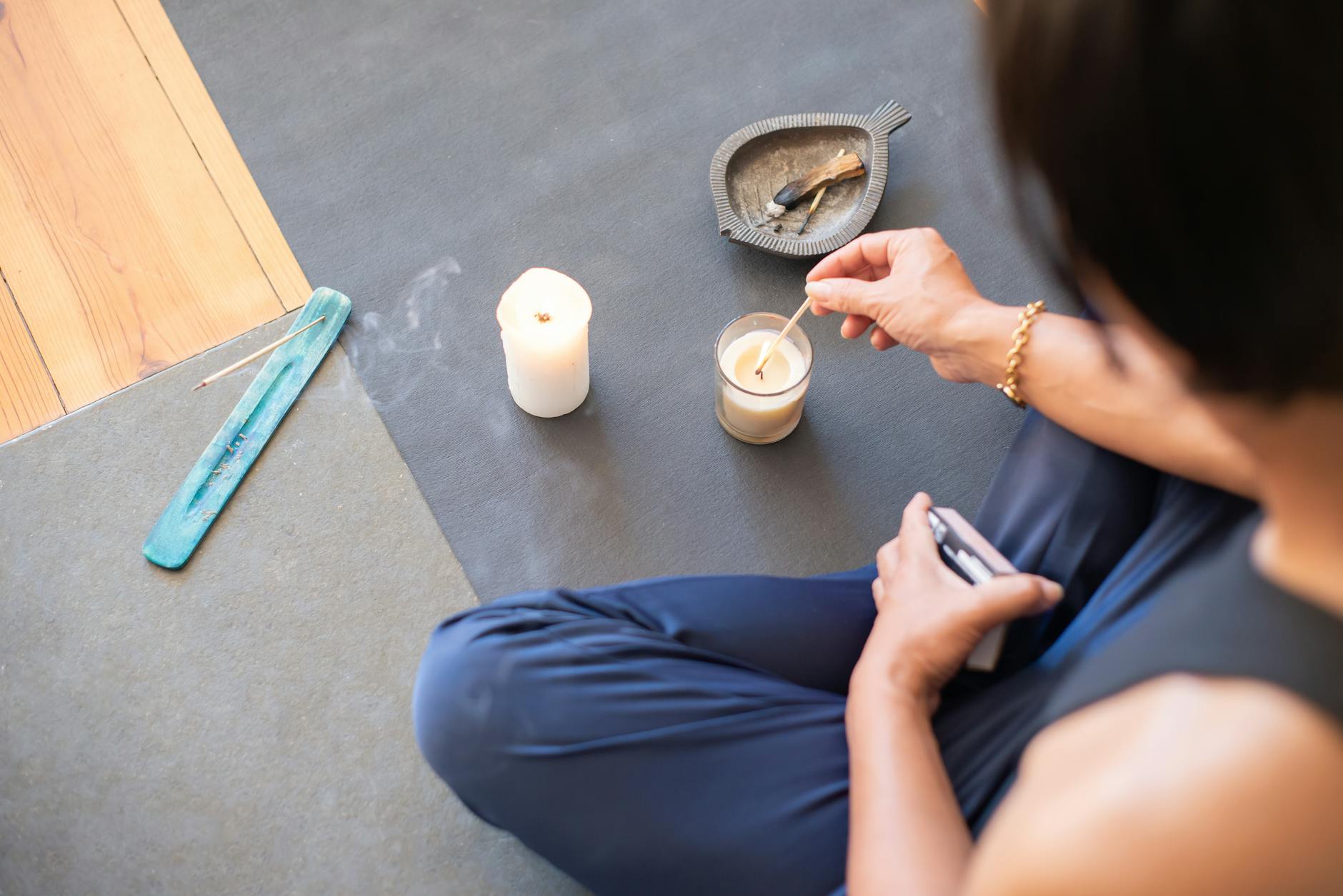 A person sitting cross-legged on a mat lighting a candle with a match, with incense and a small dish nearby