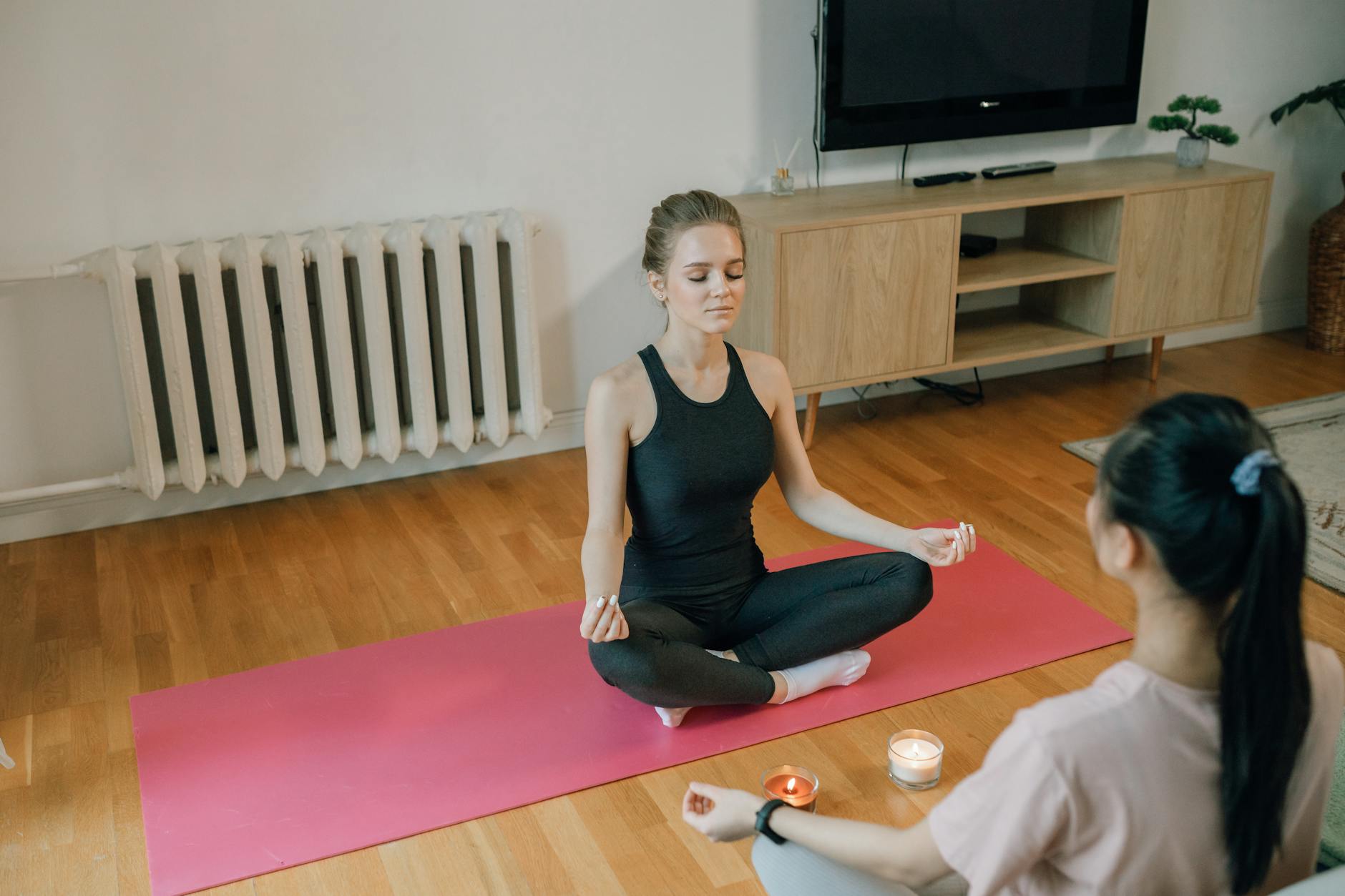 A woman sitting cross-legged on a pink mat in a living room with her eyes closed, with a second person facing her