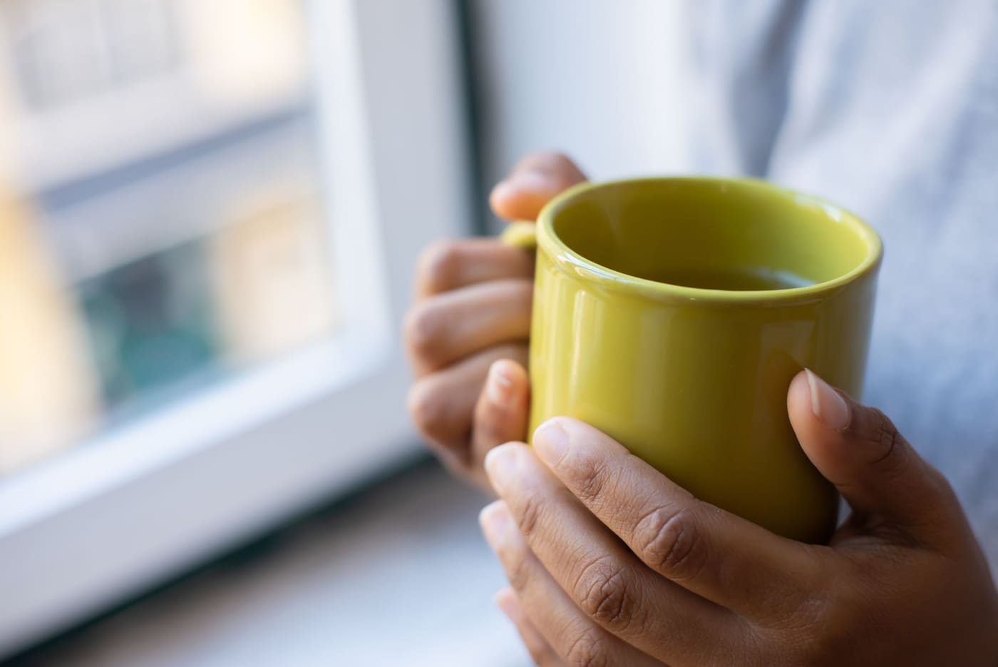 Two hands wrapped around a yellow mug beside a window