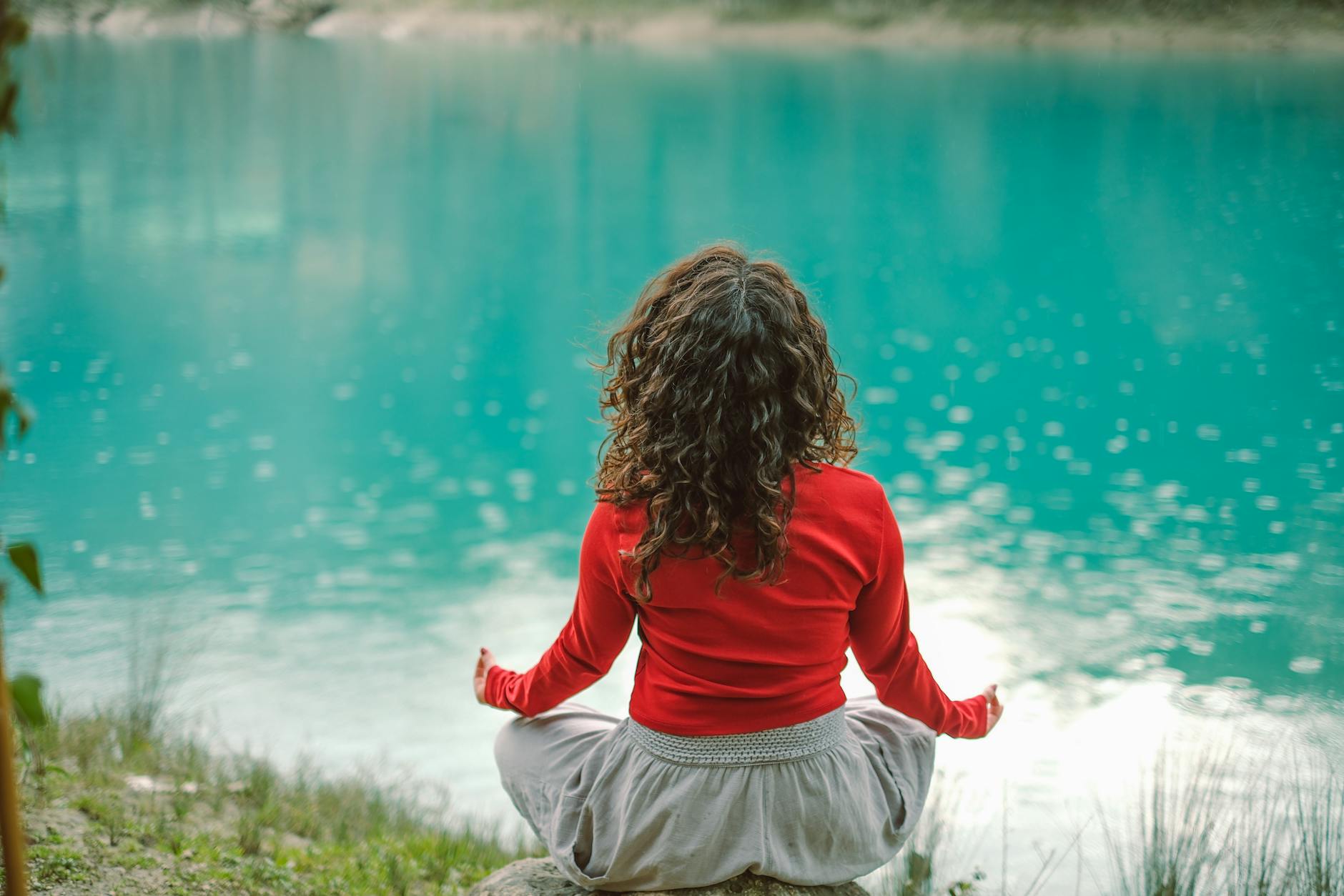 A woman with curly hair seen from behind, sitting cross-legged beside a turquoise lake